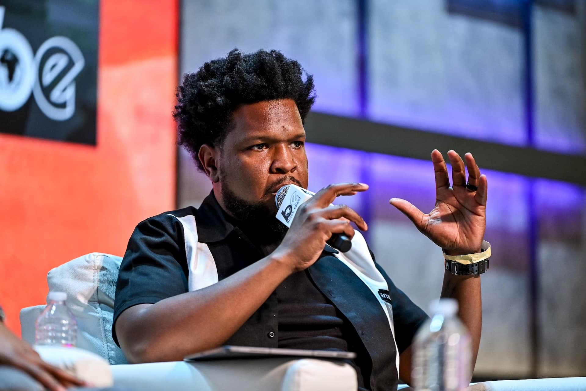 Will Lucas on a Black Effect stage with microphone, gesturing mid-sentence under blue and red lighting.