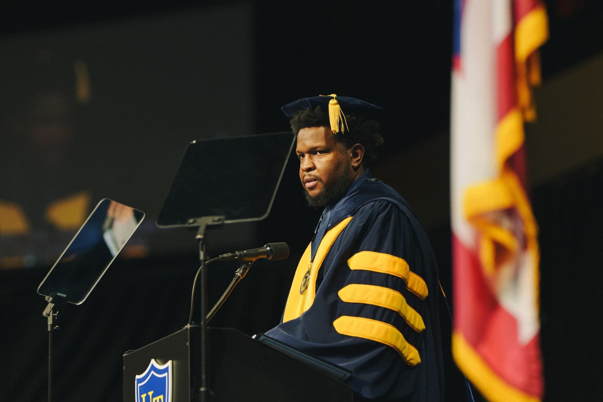 Will Lucas in doctoral cap and gown speaking from a podium with American and state flags behind him.