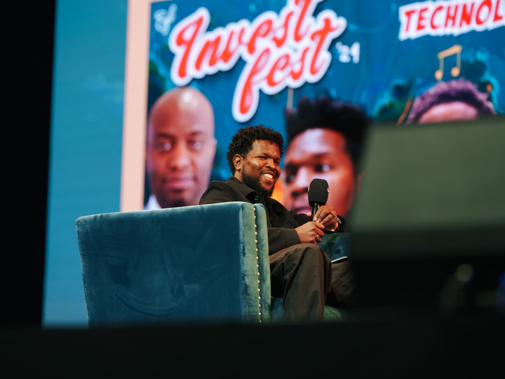 Will Lucas seated on stage with a microphone during an InvestFest panel.